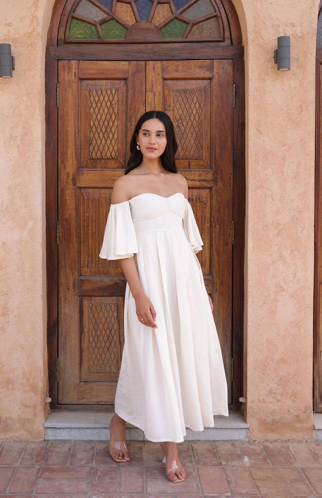 A woman stands in front of a wooden door wearing Thr3letter's Seraphina Dress in a flowing ivory color.