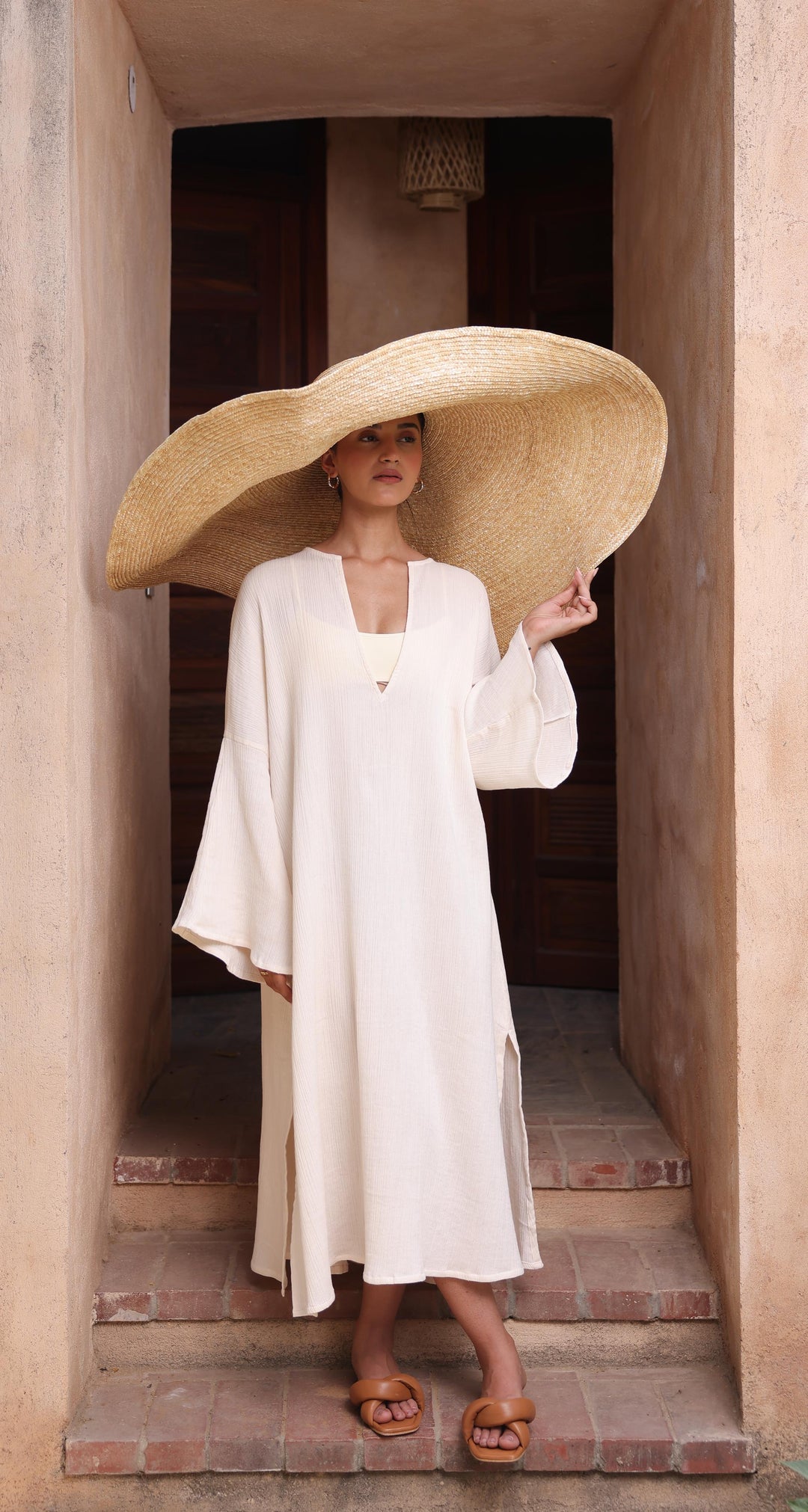 A woman stands in a doorway wearing Thr3letter's flowing Kiaan Kaftan in a soft, neutral almond color.