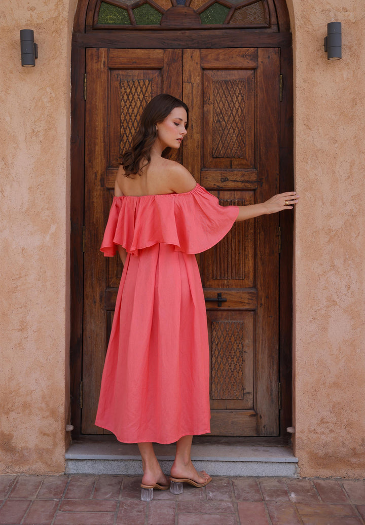 A model wears Thr3letter's Seraphina Dress in a vibrant salmon tone, complementing the architectural backdrop.