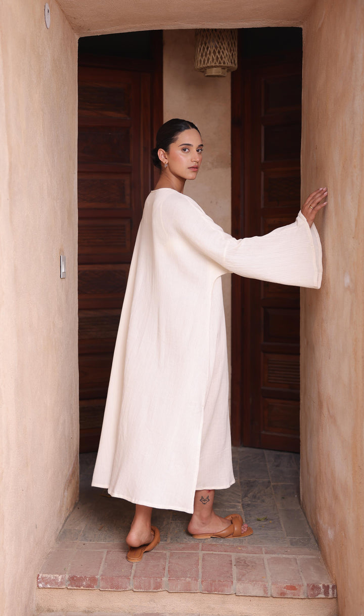 A woman models the Thr3letter Kiaan Kaftan in an understated almond tone, framed by a wooden doorway.