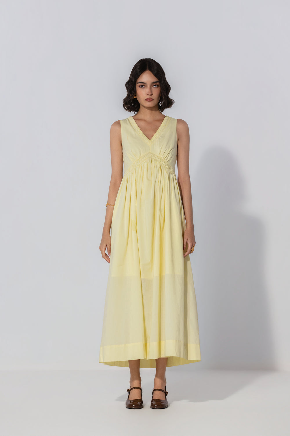 Women's pastel yellow sleeveless V-neck midi dress with an empire waist, gathered bodice, and flowing A-line skirt styled in a studio.